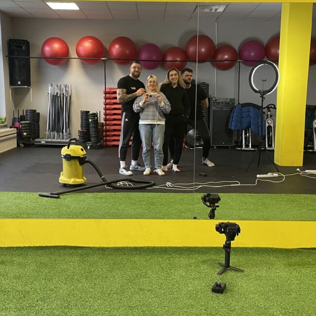 grupa ludzi: dwie kobiety. dwóch mężczyzn, stoją przed lustrem w salce treningowej siłowni RMG GYM w Kielcach i robią sobie zdjęcie pamiątkowe po nagrywkach dla sieci siłowni RMG GYM
